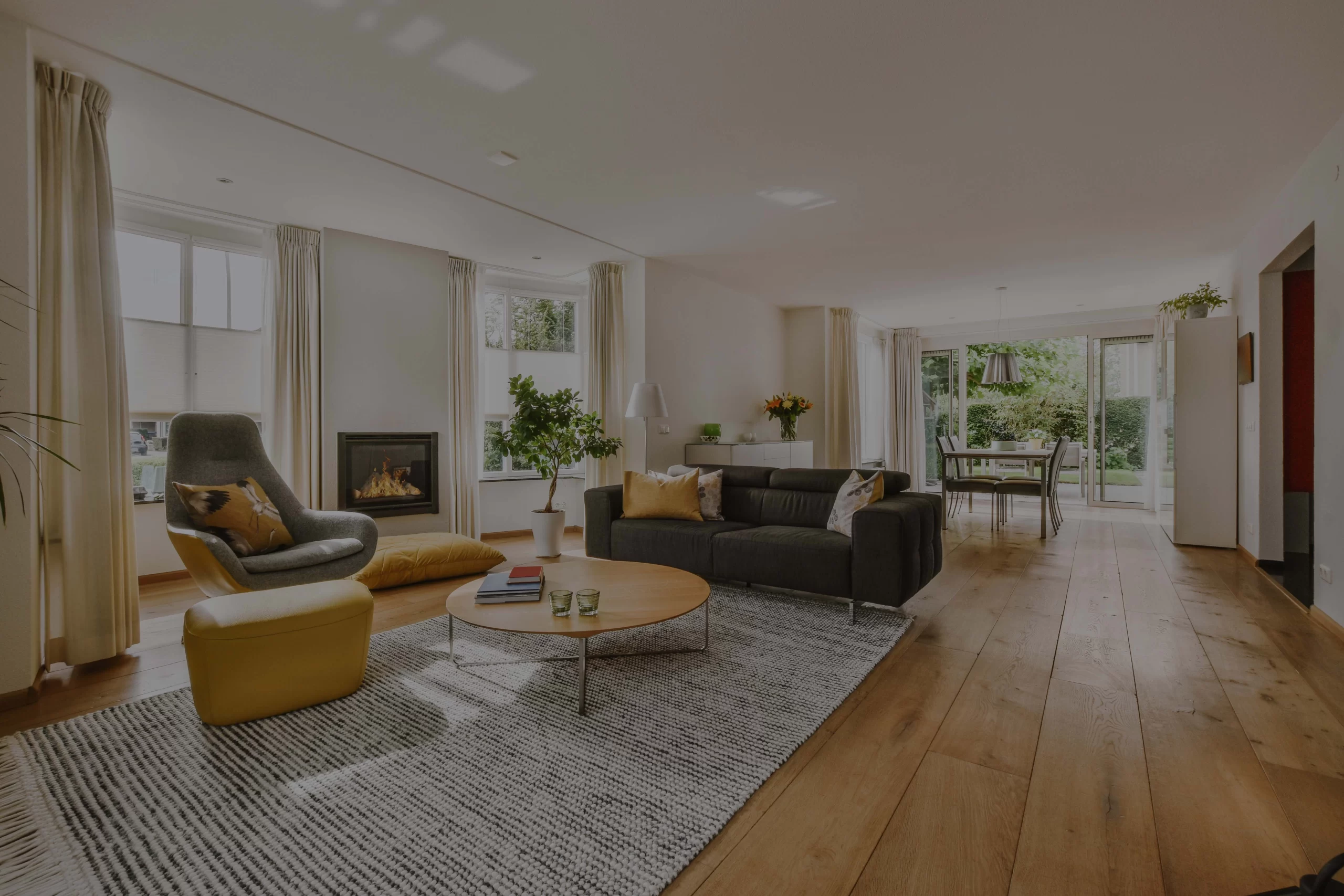 Modern living room with wooden floors, featuring a gray armchair and yellow ottoman near a cozy fireplace, dark sofa, large rug, and dining area with garden view.
